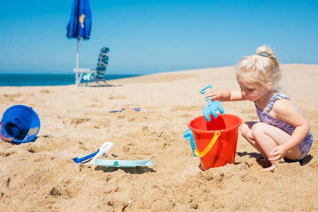 child-playing-in-sand-with-red-bucket-and-blue-shovel-on-sunny-beach