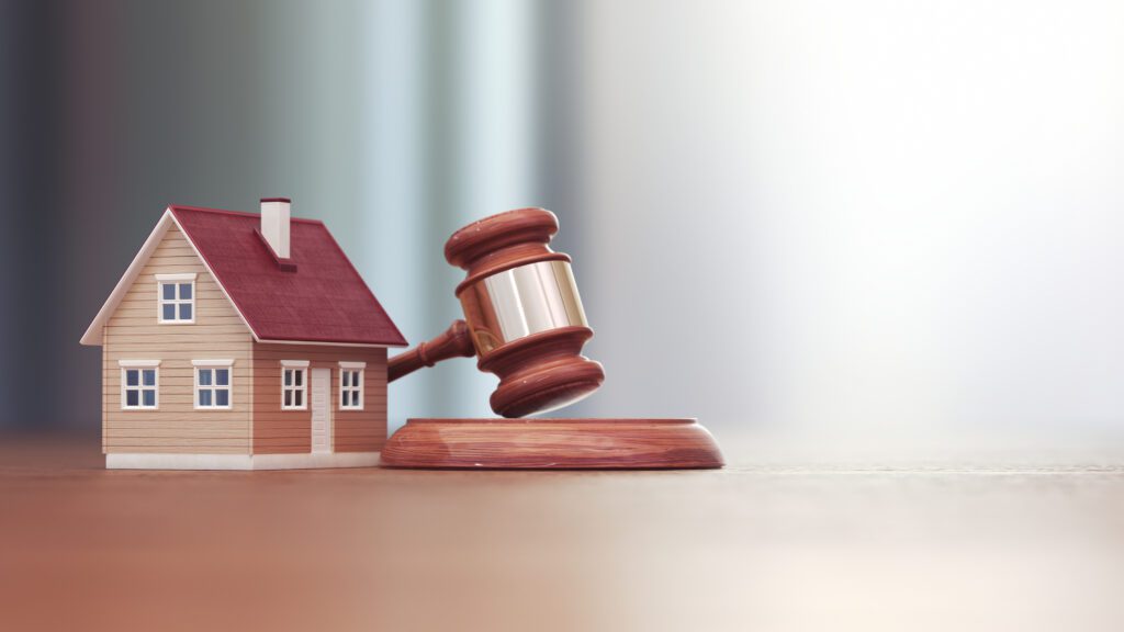 A small wooden house model with a red roof sits on a smooth surface beside a wooden gavel