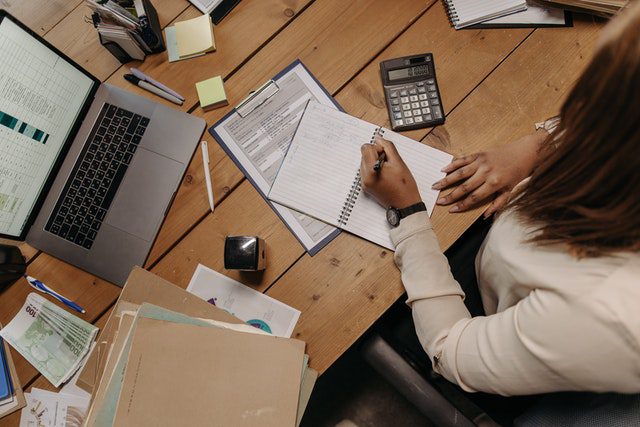person-writing-notes-in-notebook-on-desk-with-laptop-calculator-and-documents