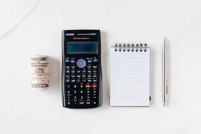 rolled-stack-of-dollar-bills-a-scientific-calculator-a-notepad-and-a-pen-on-a-white-surface